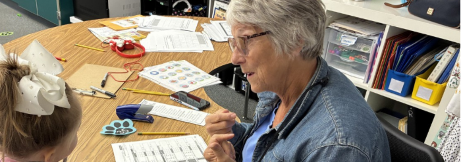 A woman in a denim jacket engaged in a conversation with a child at a table.