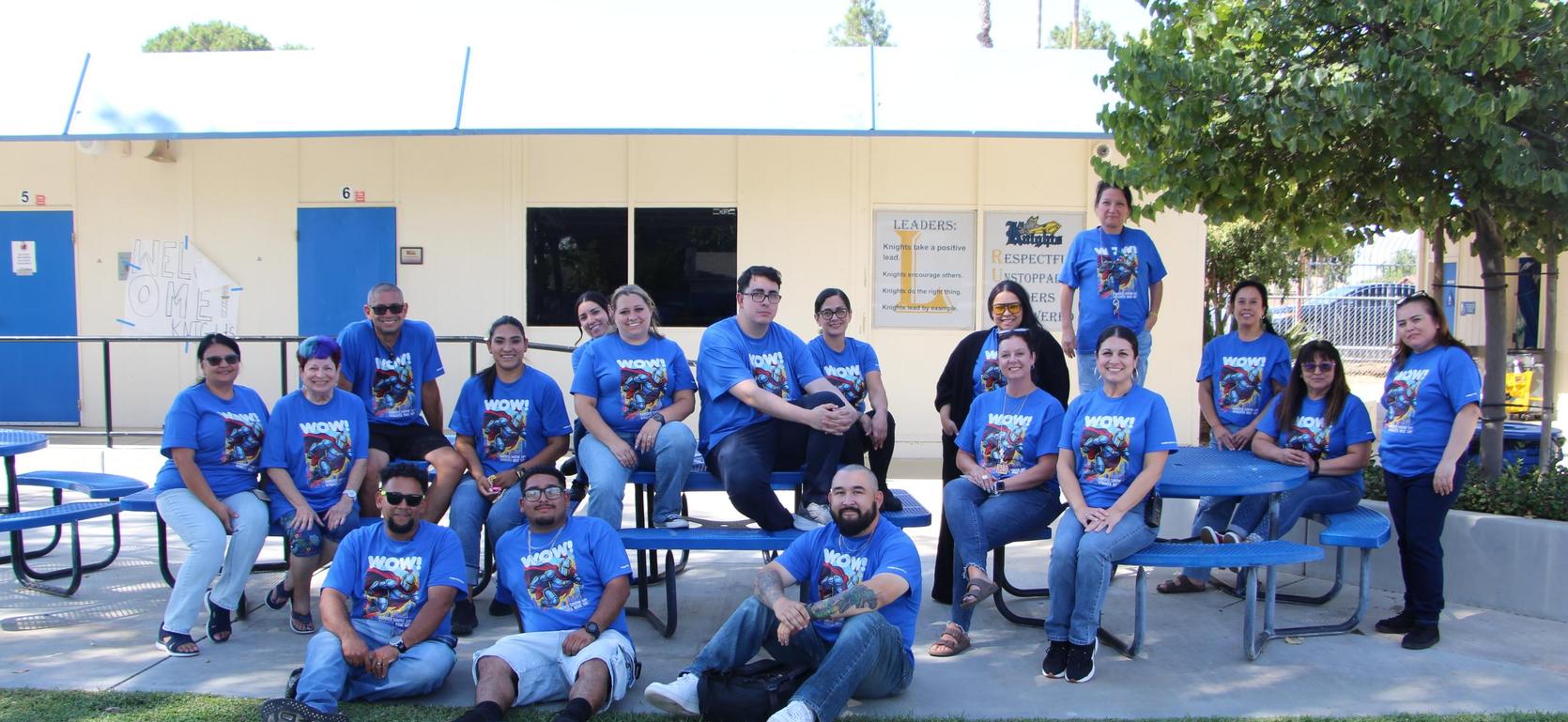 Group of people in matching blue t-shirts sitting and standing around picnic tables.