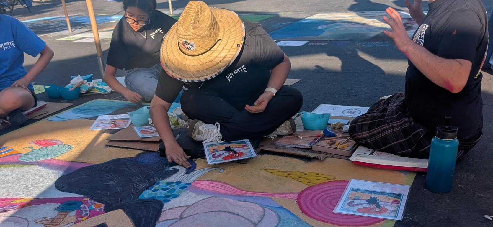 Artists color a large chalk mural on the ground, surrounded by tools and sketches.