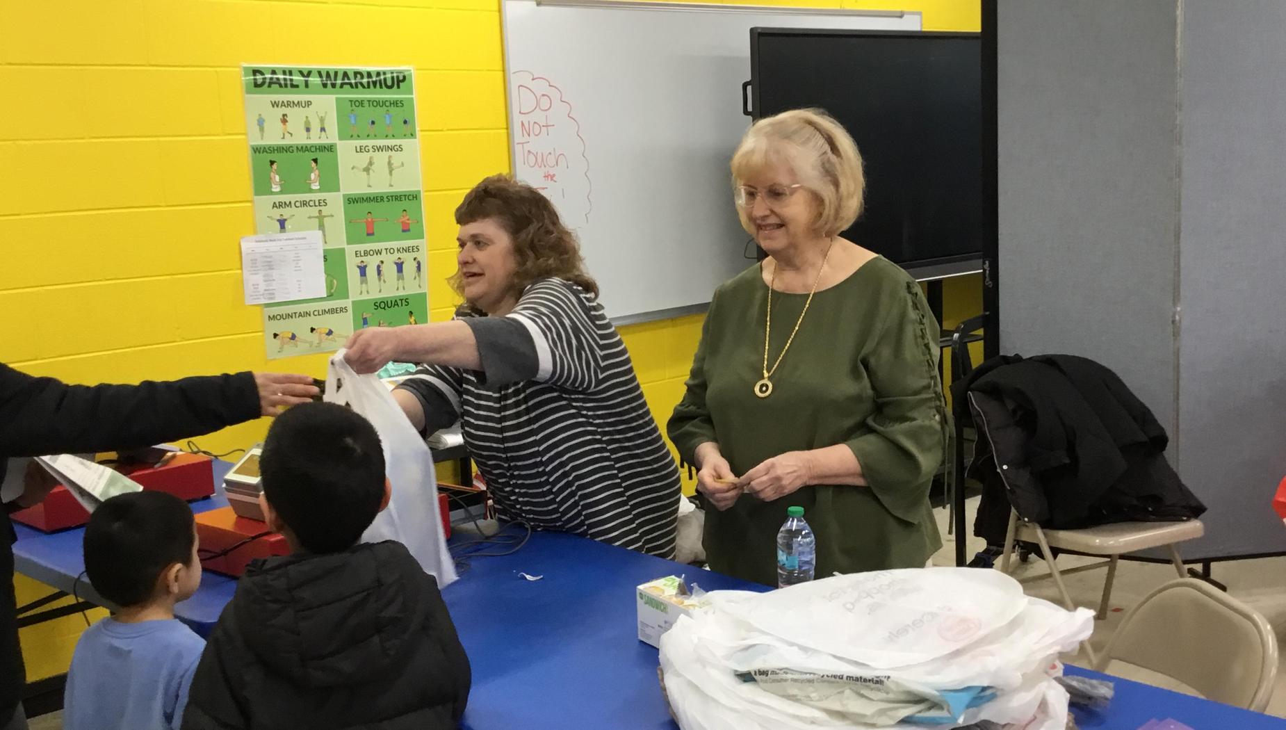 Two women assist children with bags at a table in a vibrant classroom setting.