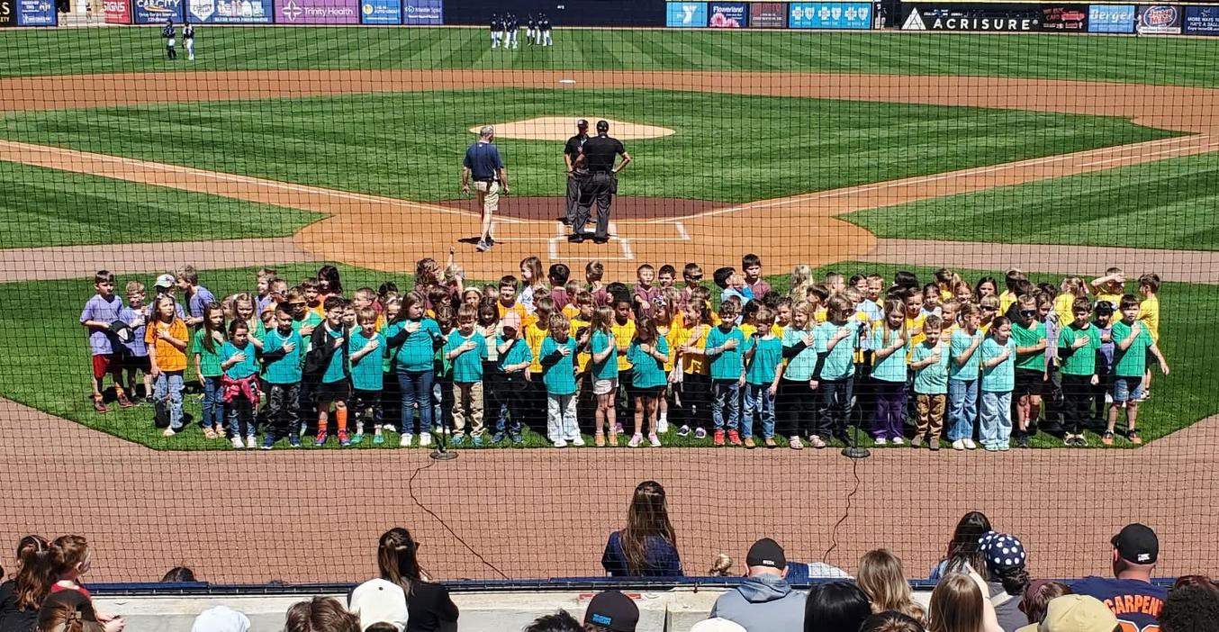 Children in colorful shirts gather on a baseball field for a performance.