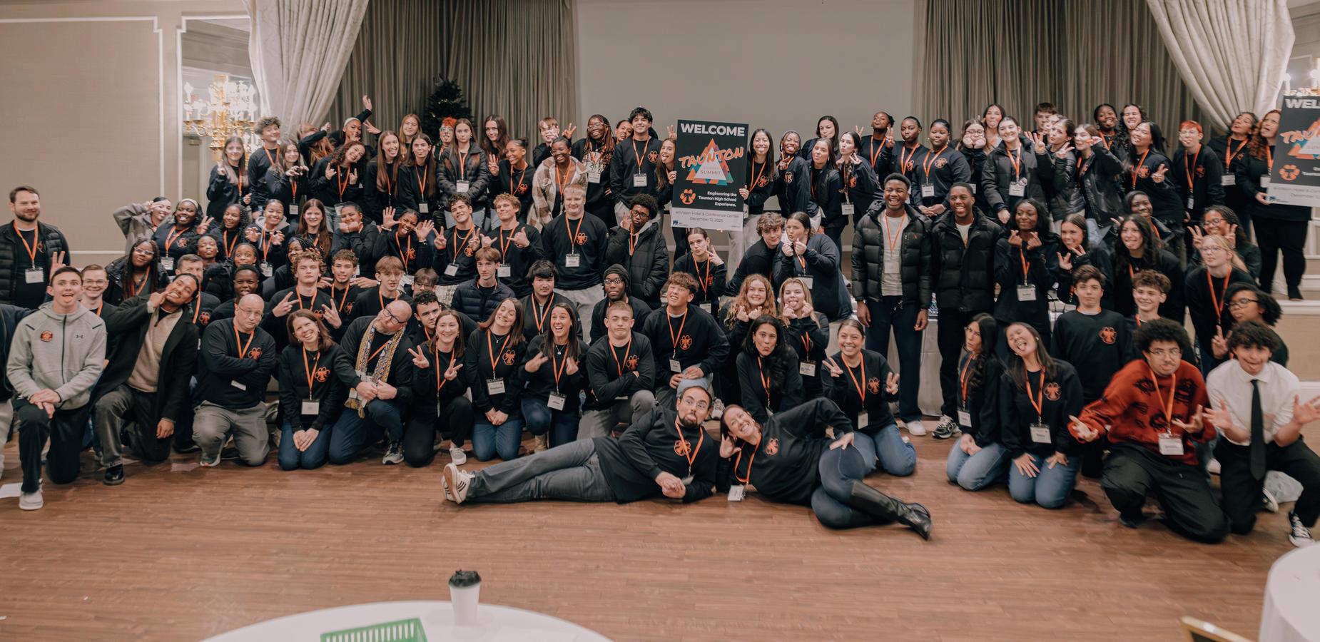 A large group photo of diverse students and adults posing in a ballroom for the Taunton Student Summit. The group is wearing matching black shirts with orange logos and lanyards, with many smiling, making hand gestures, and posing playfully around a sign that reads "Welcome Taunton Student Summit."
