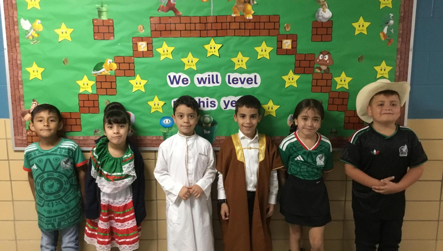 Group of children in festive outfits standing in front of a colorful bulletin board.