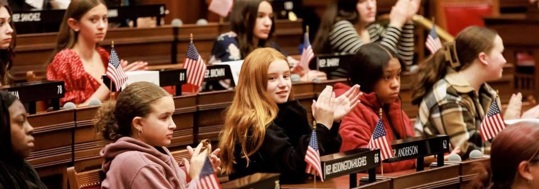 Young girls sitting in a legislative chamber, clapping with small flags on their desks.