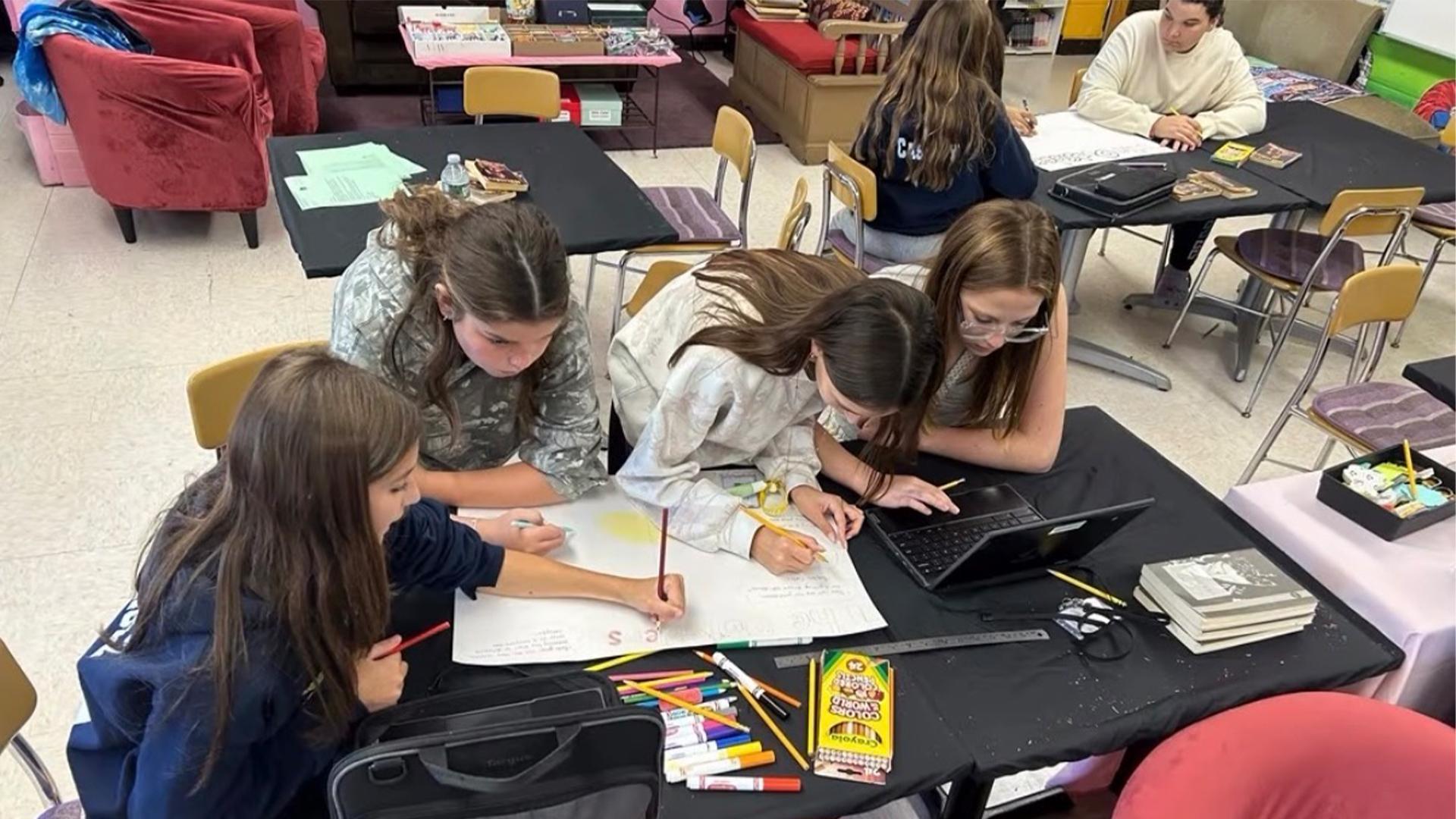 Four girls collaborating on a project at a table, with notebooks and art supplies around them.
