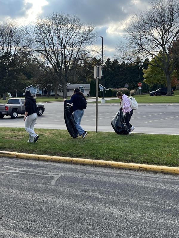 Three individuals walking on grass carrying bags in a parking area.