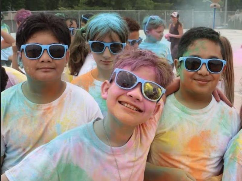 Group of children smiling, wearing colorful shirts and sunglasses, at a festive event.