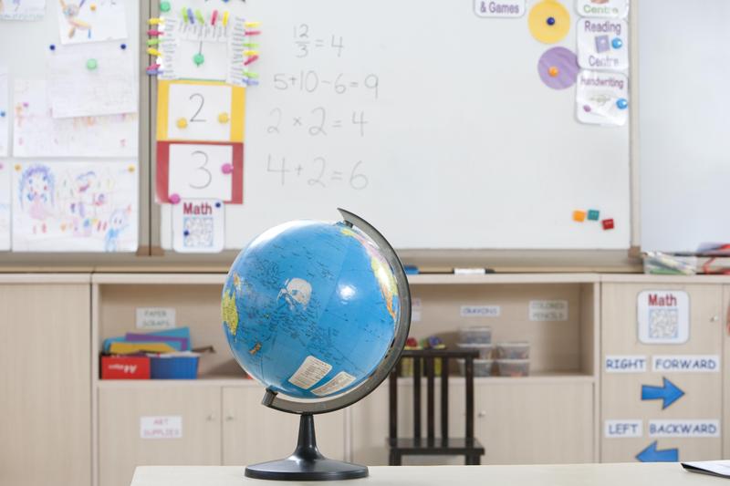 A classroom setting presents an inviting tableau of learning. A blue globe with continents stands prominently on a light table, acting as a visual centerpiece. In the background, a whiteboard displays colorful designs and simple math problems. Labels in clear fonts indicate different learning centers. The overall mood is one of lighthearted engagement, making this image ideal for use in educational materials, school websites, or projects related to geography, education, and early childhood development.