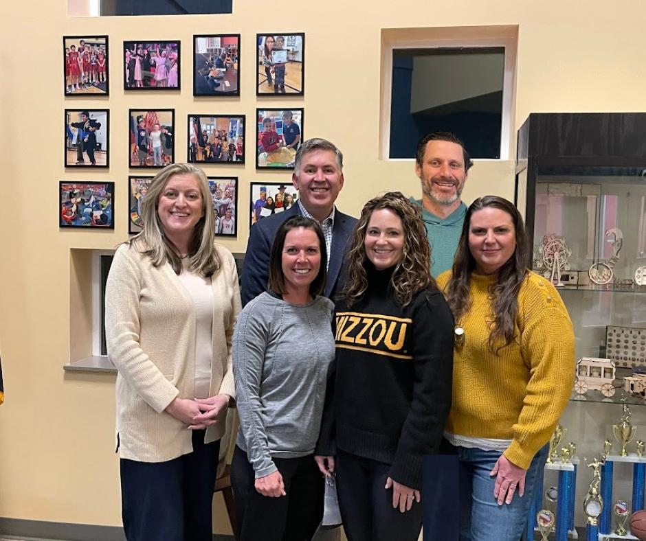 Photo of six board members standing in front of flags and trophy case.