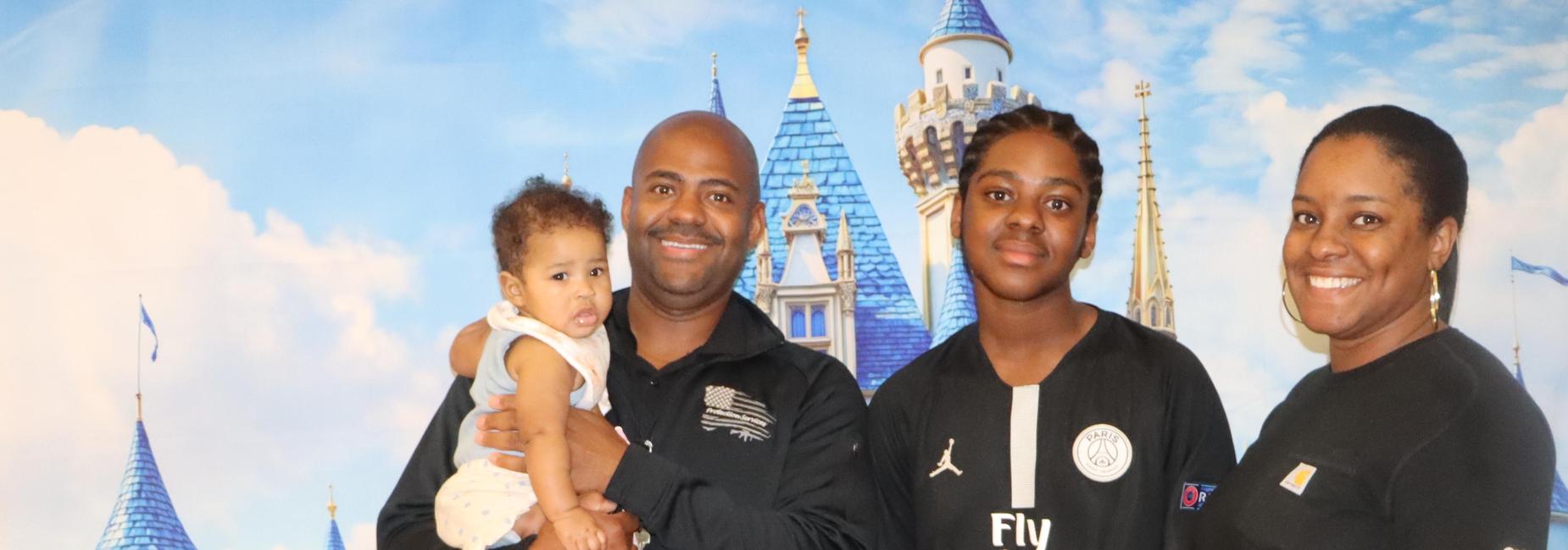 Family portrait with a father holding a baby, an older child, and a mother in front of a castle backdrop.