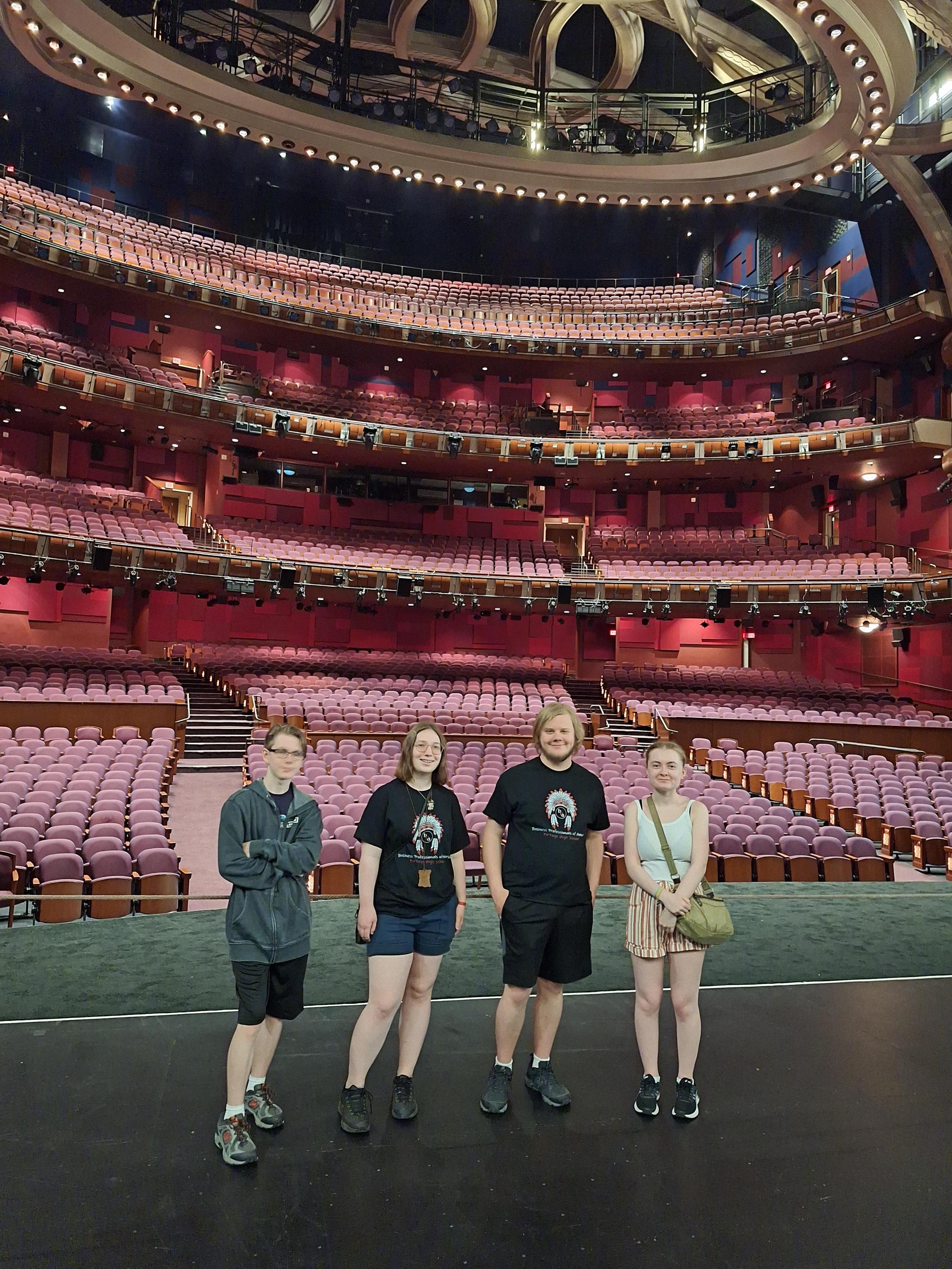 Four individuals standing on stage in a theater with empty seats.