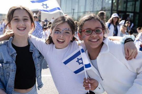 Three smiling girls holding Israeli flags at a festive event.