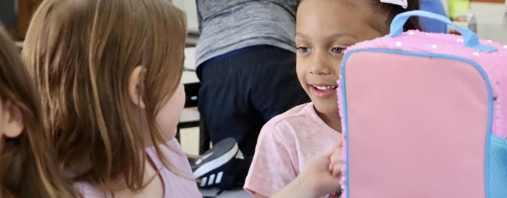 Two young girls smiling and chatting with each other in a classroom.
