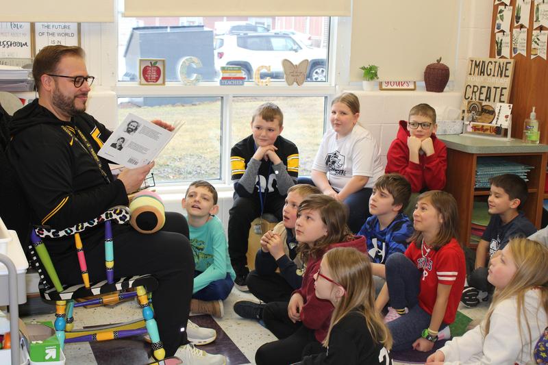 students excitedly looking at principal who is sitting in a rocking chair reading to them