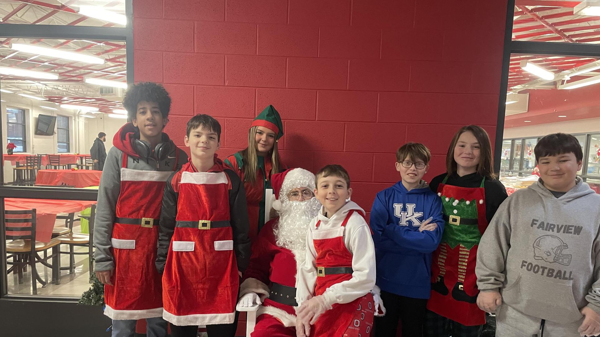 A group of children and an elf pose with Santa in festive attire.