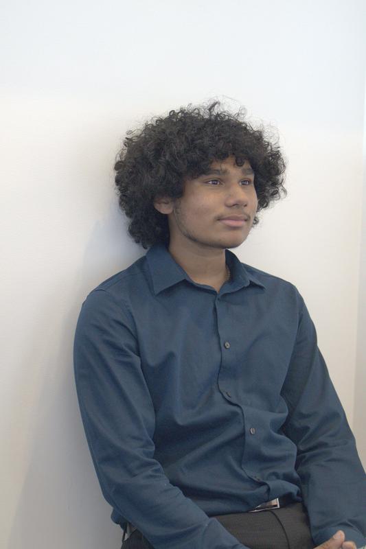 Young man with curly hair sitting against a wall, wearing a blue shirt.