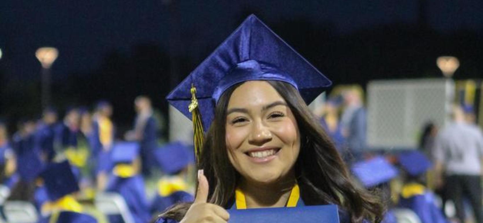 Smiling graduate in cap and gown giving a thumbs up holding a diploma.
