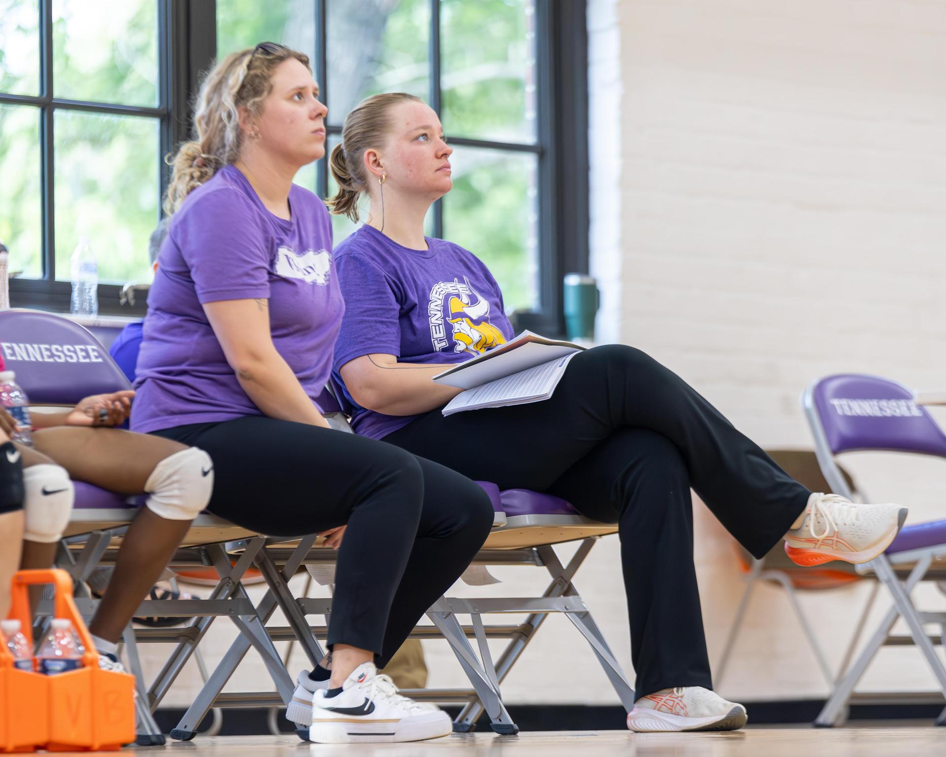 Two women sit on bleachers, both wearing purple shirts, watching the game.
