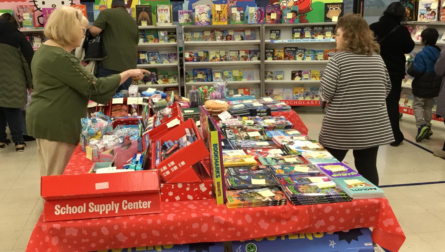 A school supply table filled with books and items, with adults interacting around it.