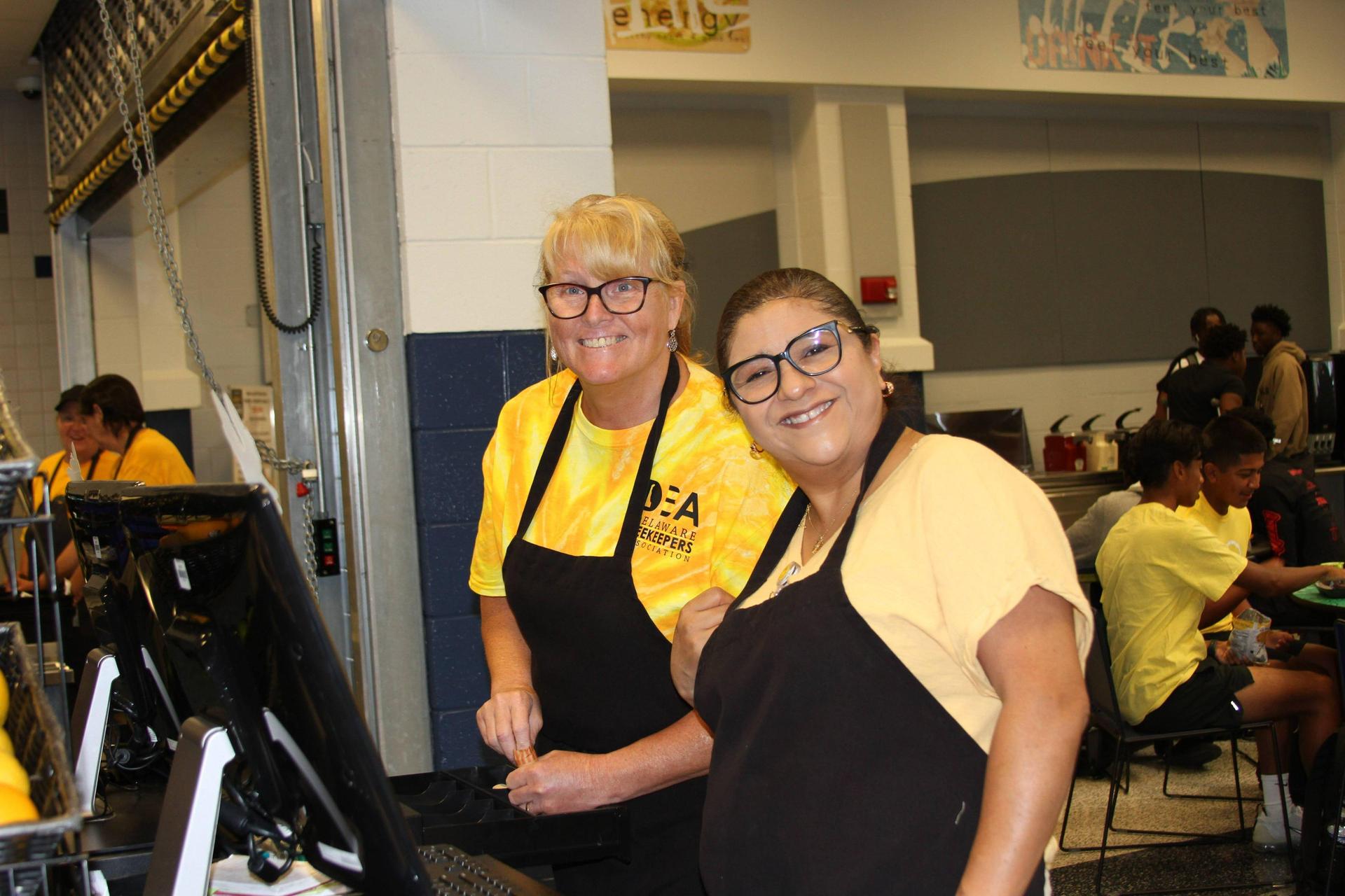 Two smiling cafeteria staff in yellow shirts standing behind serving stations.