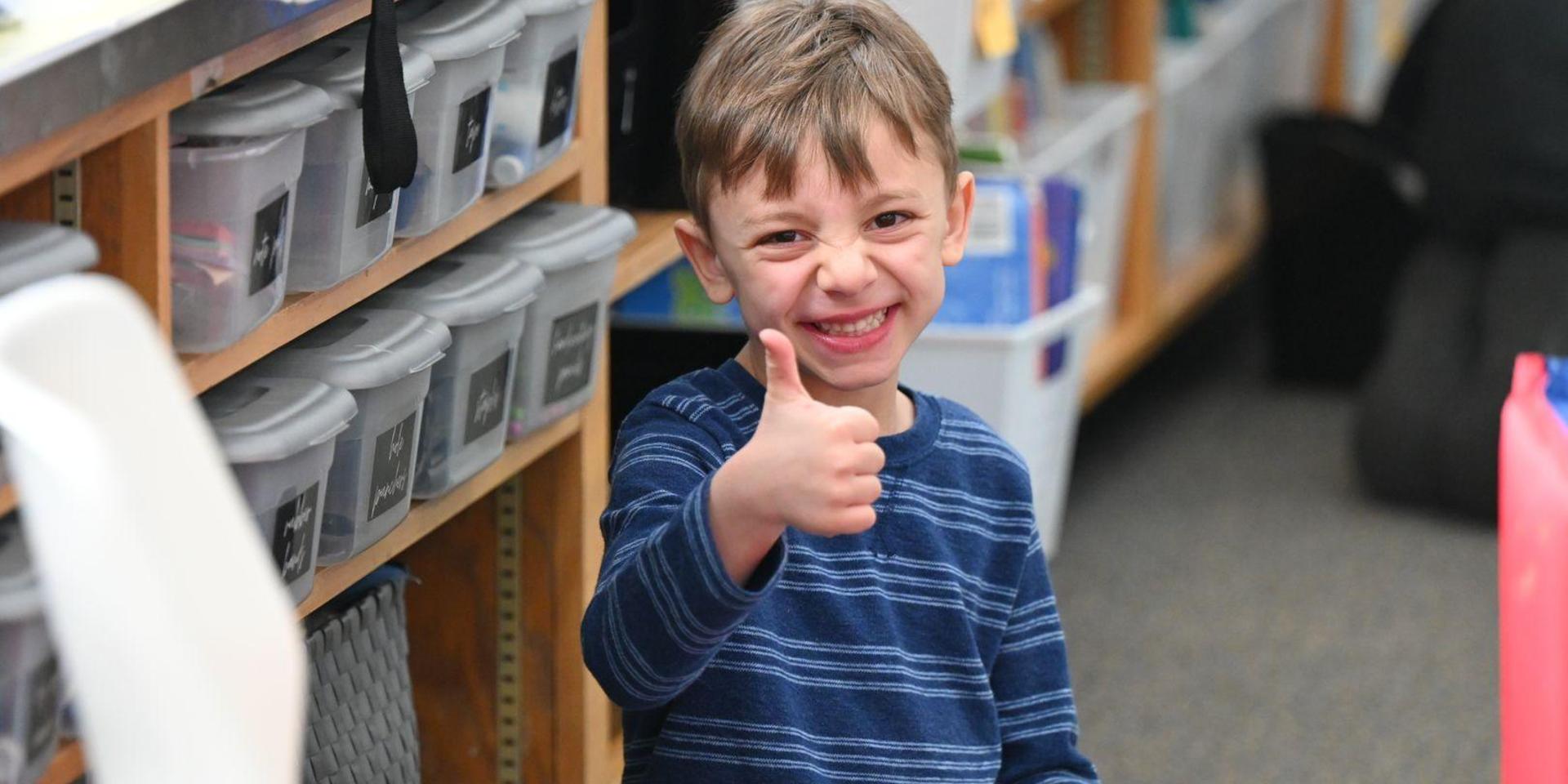 Young boy giving a thumbs up while sitting in a classroom.
