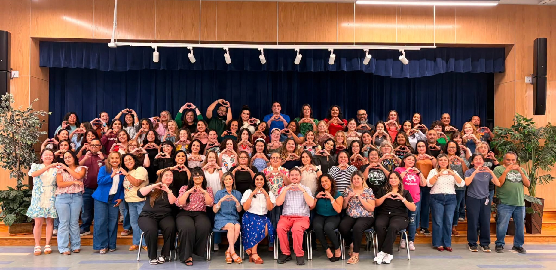 Large group of people making heart shapes with their hands in a school auditorium.