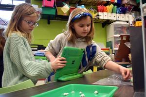 Two girls collaborate on a project, with craft supplies and a tablet on the table.