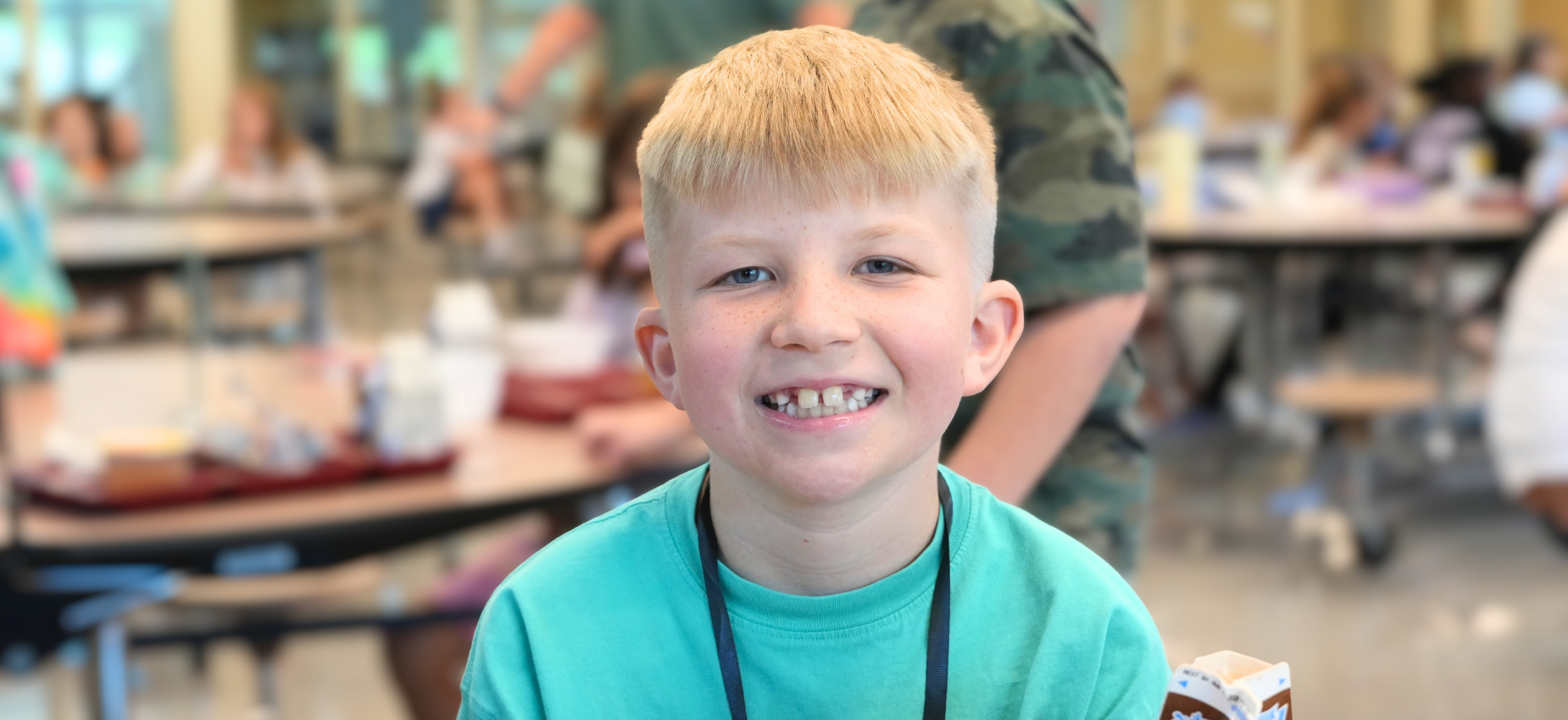 Smiling young boy with blonde hair in a busy school cafeteria.
