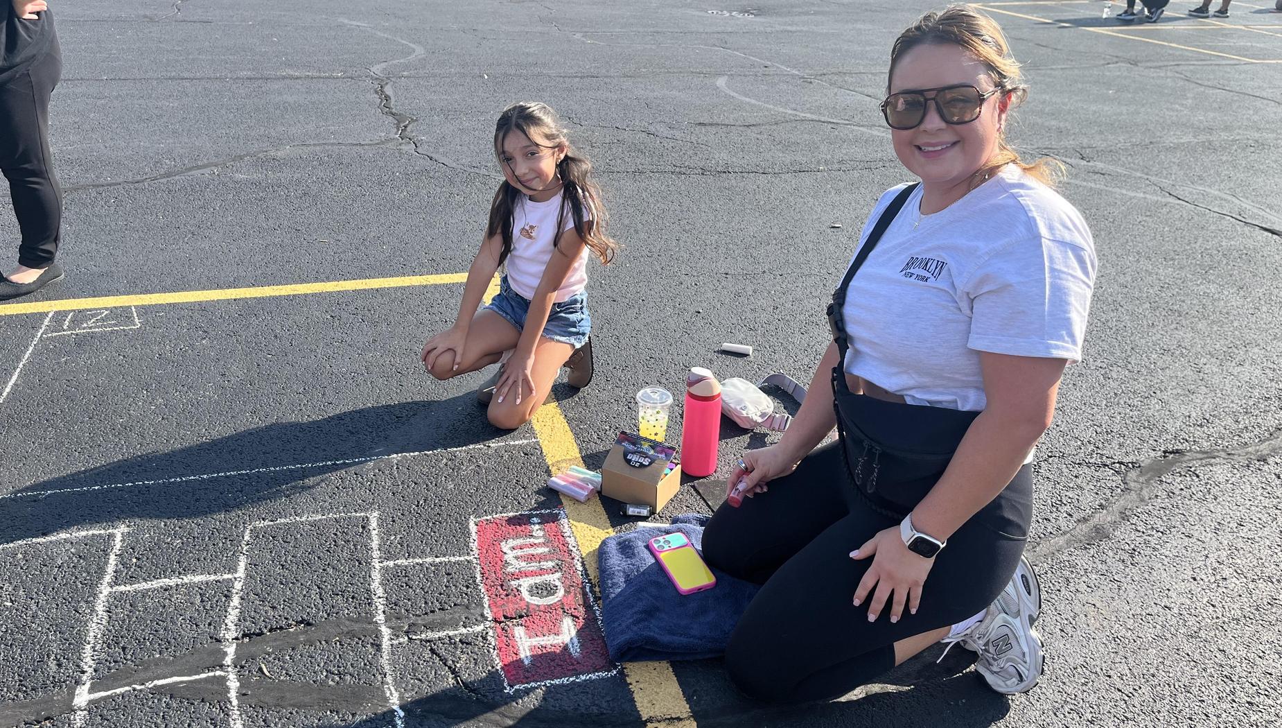 A girl and a woman kneeling next to chalk art on pavement, both smiling at the camera.