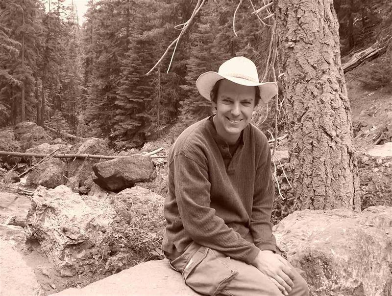 Smiling man in a hat sitting on a rock near a forested area.