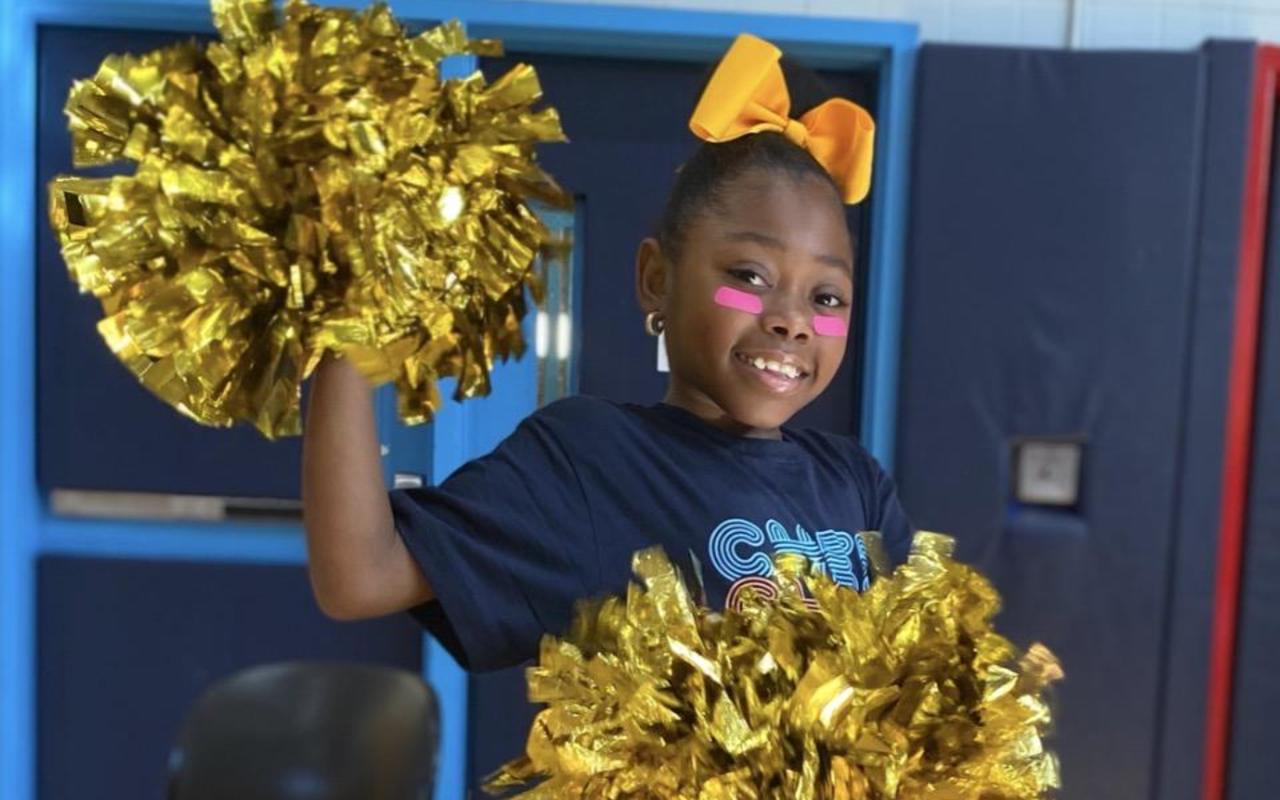Child holding gold pom-poms, wearing a blue shirt in a gym setting.