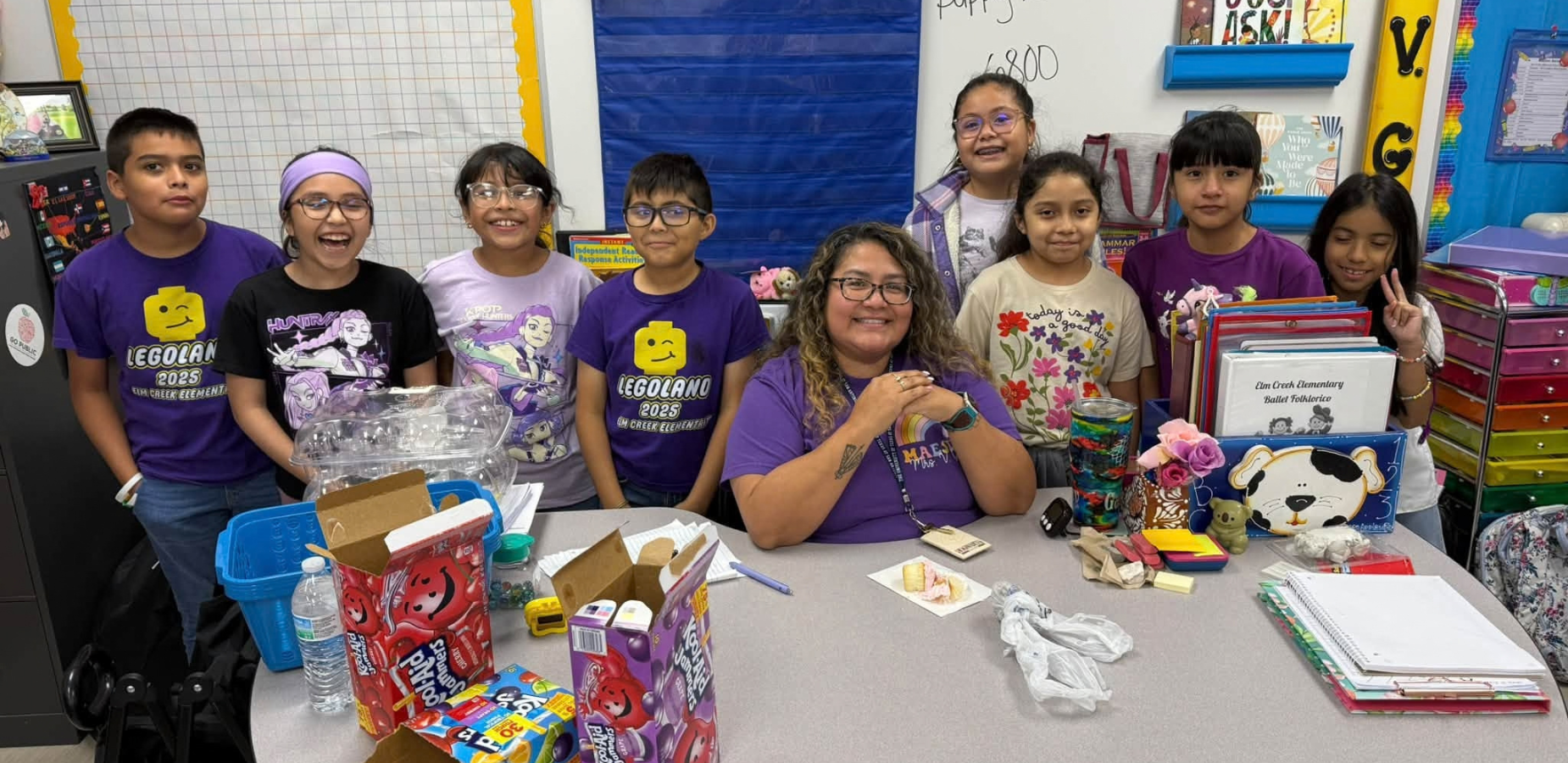 Group of children and a teacher smiling at a classroom table with various colorful items.
