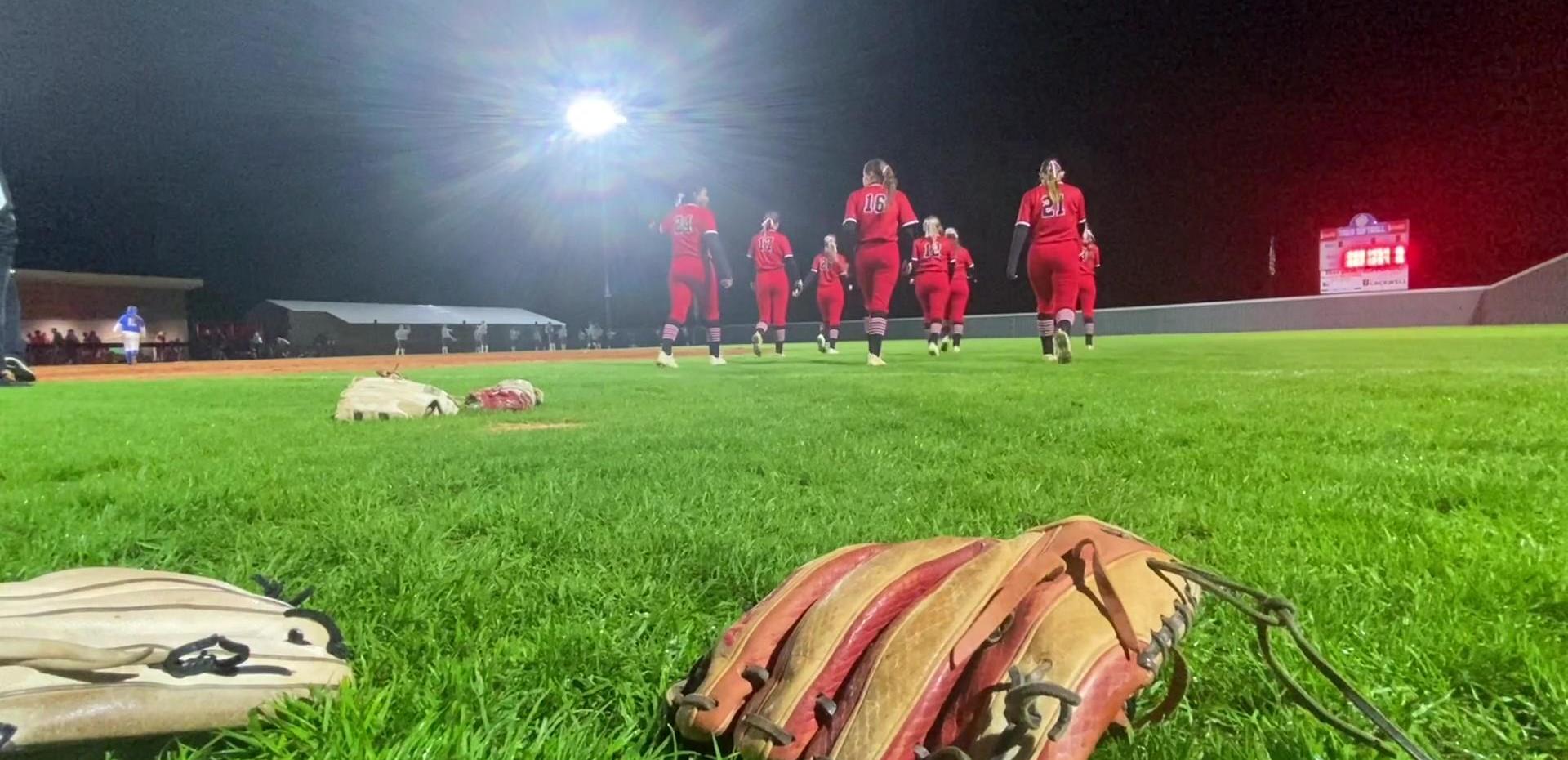 Softball players in red uniforms running towards the field under stadium lights.