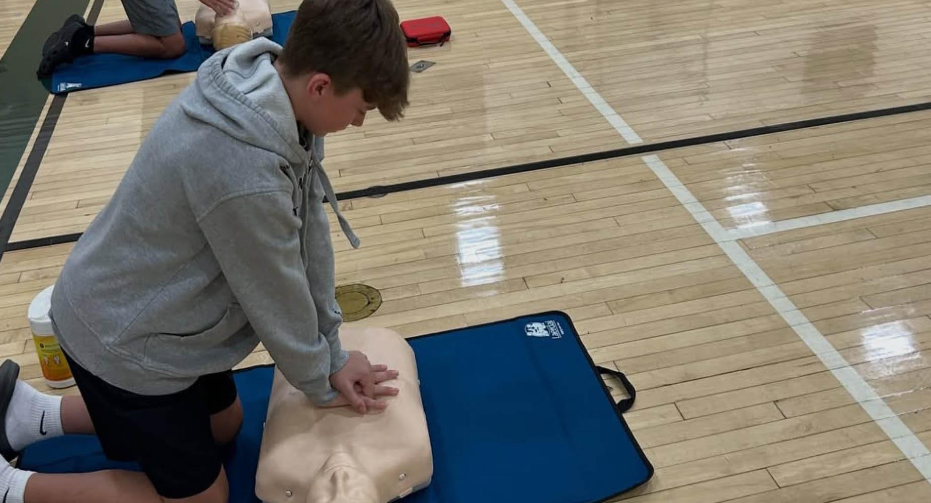 Student performing CPR on a training mannequin in a gym.
