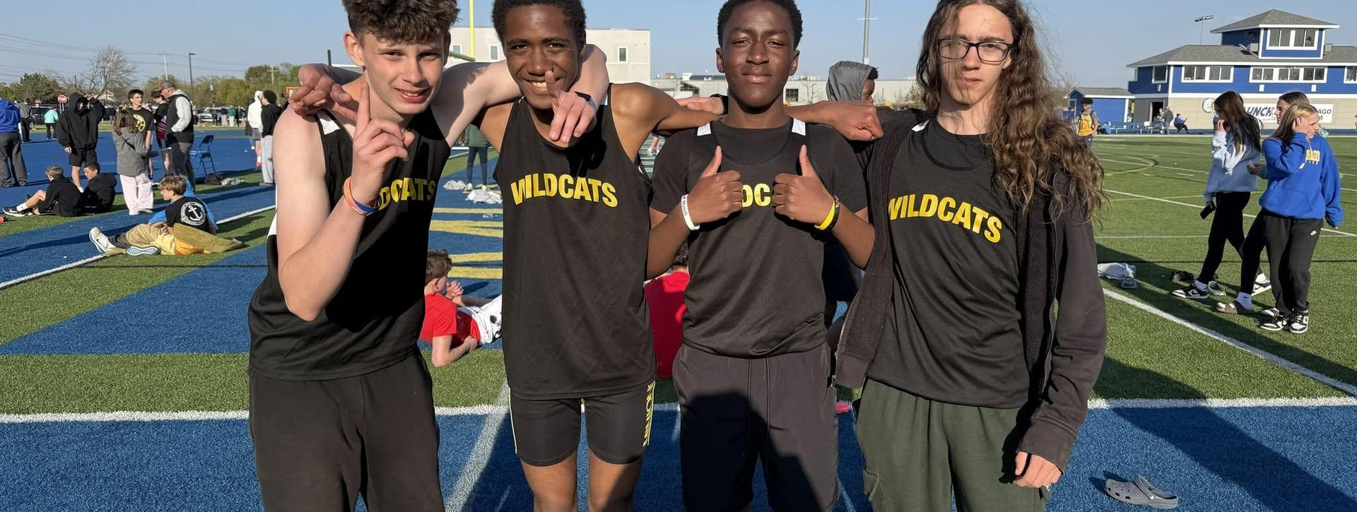 Four male athletes in black jerseys with 'WILDCATS' posing on a sports field.