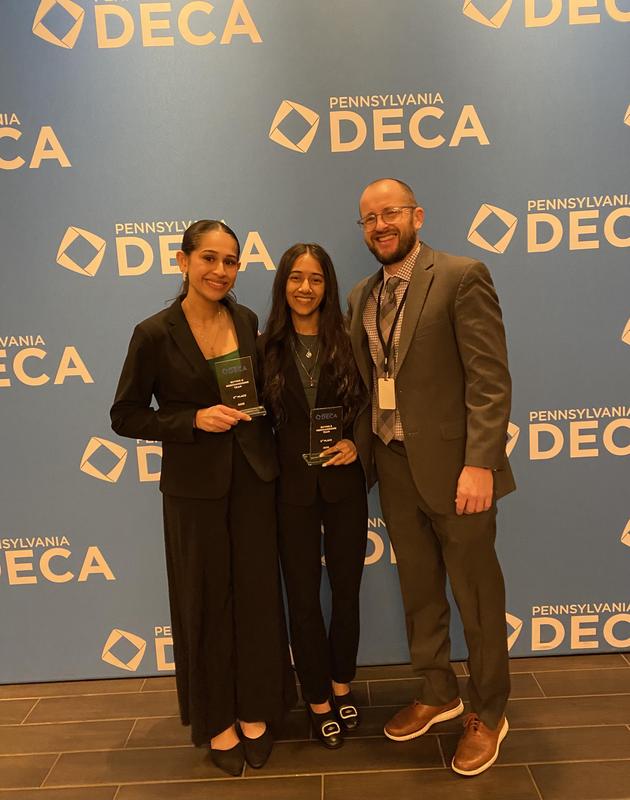 Image of 2 students and their advisor standing in front of a backdrop that says DECA