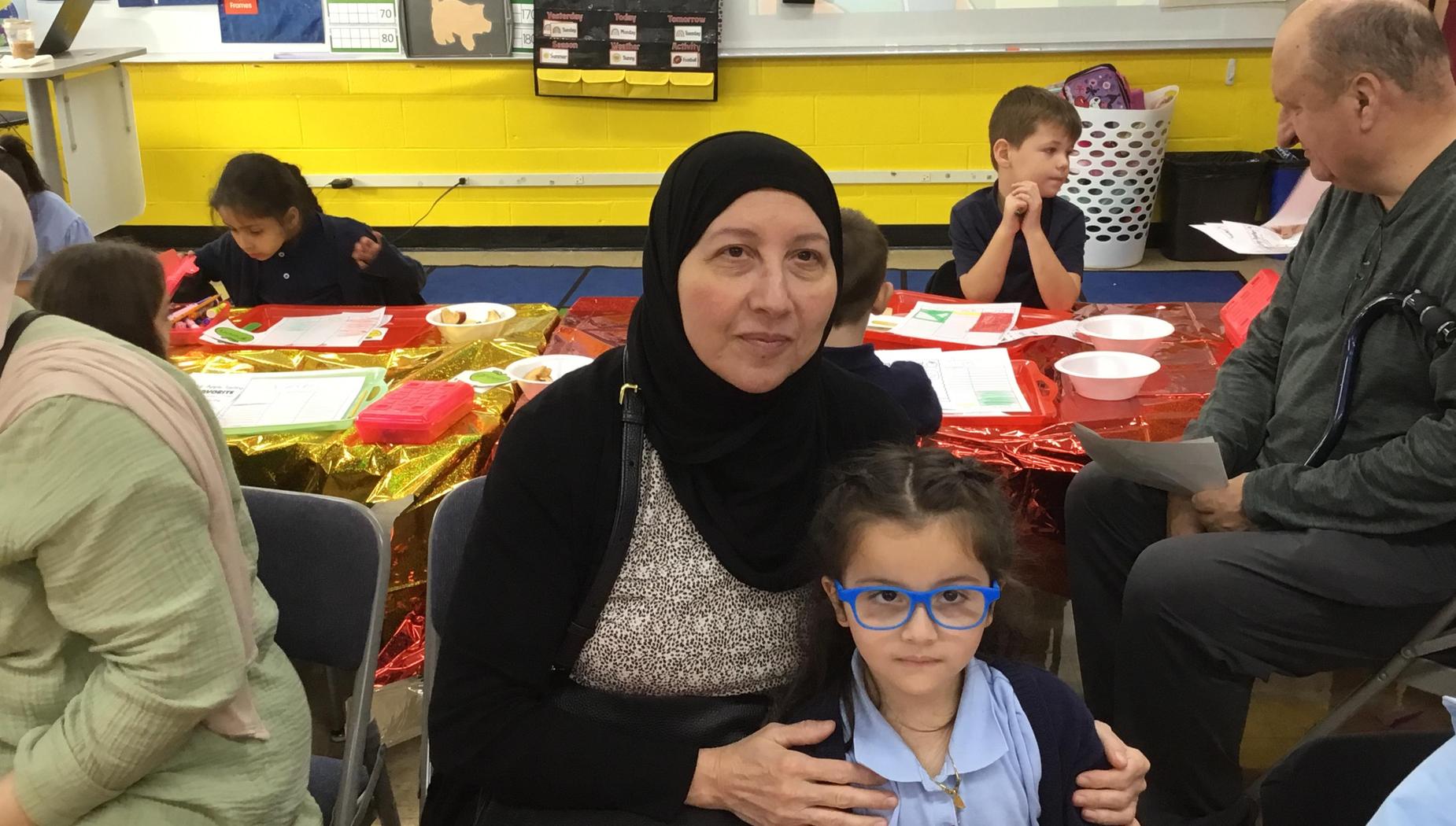 A woman in a black hijab sits with a girl wearing blue glasses in a classroom.