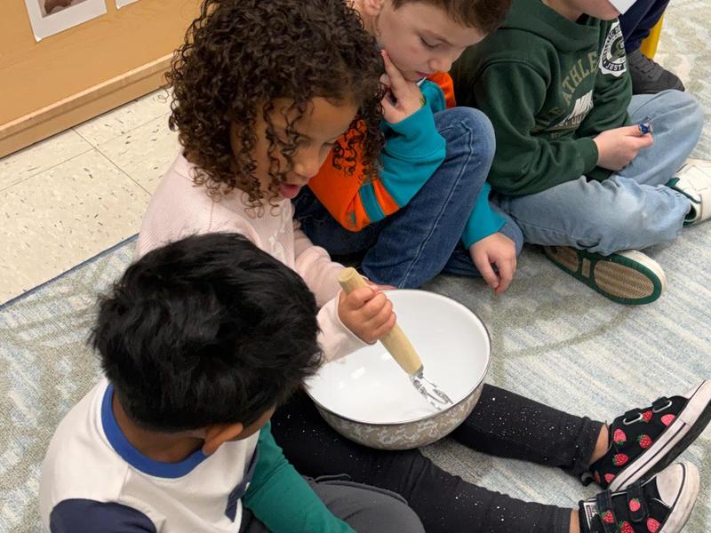 Children sitting together on a mat, one stirring a bowl, focused on an activity.