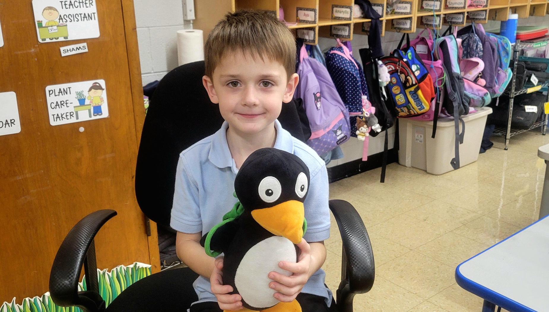 A boy sitting in a classroom holding a stuffed penguin toy.