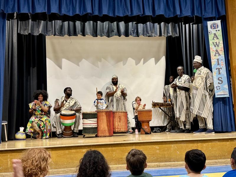 African musicians are dressed in traditional African garments and playing instruments from Africa while on stage with two small children playing drums.