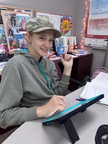 Student working at a desk with a tablet, smiling and wearing a cap.