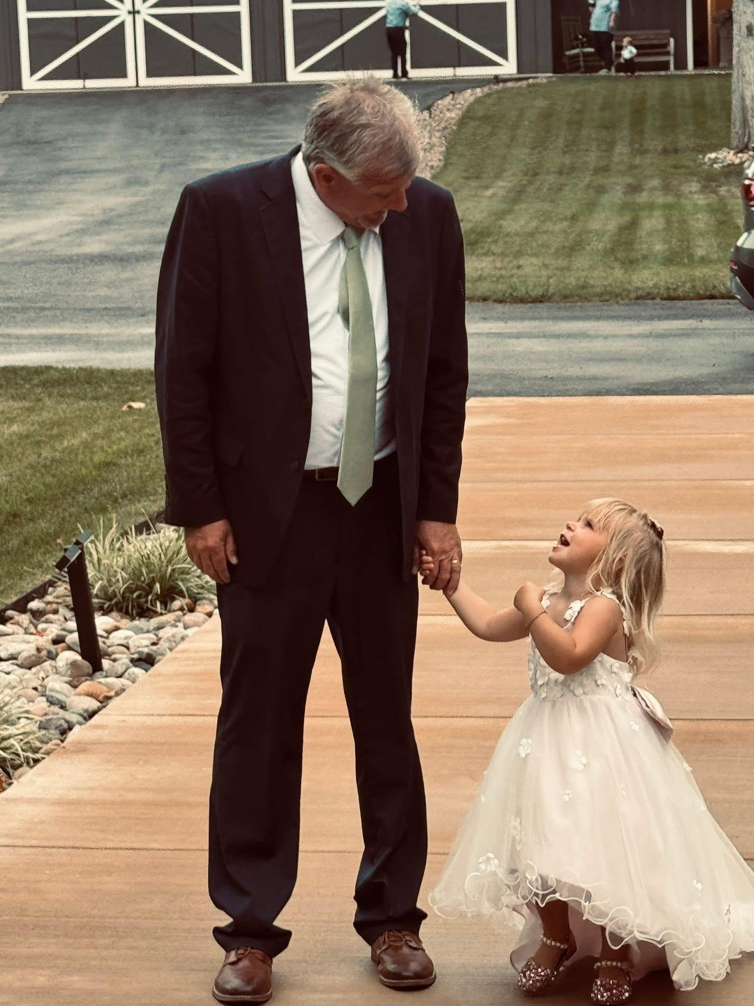 A girl in a white dress stands with a man in a suit, both smiling at each other.