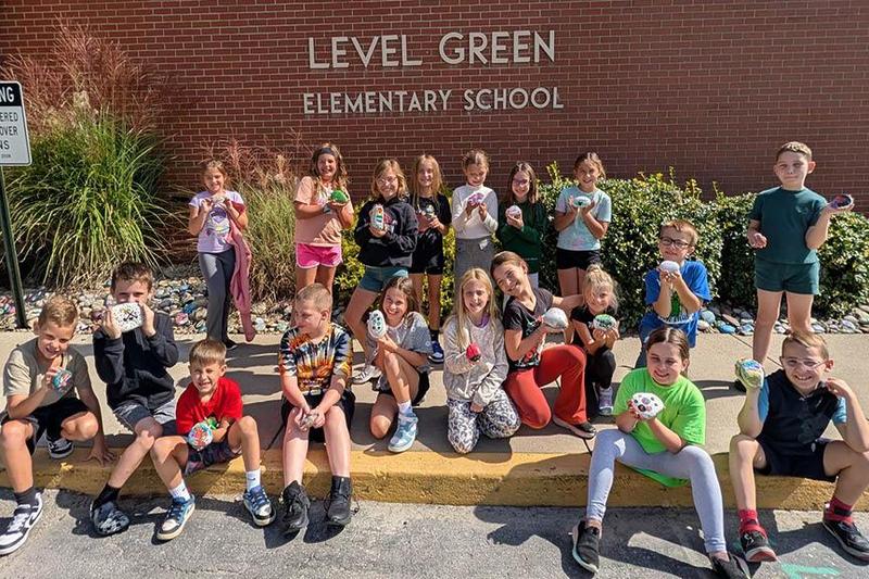 a large group of students stands outside a school and each one is holding a rock