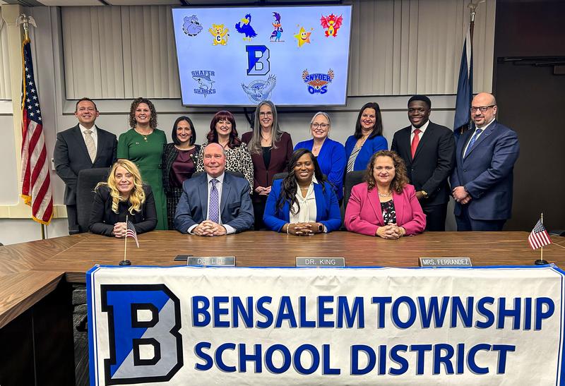 Image of 13 woman and men 4 sitting at the meeting room table and 9 standing behind them with text that says Bensalem Township School District