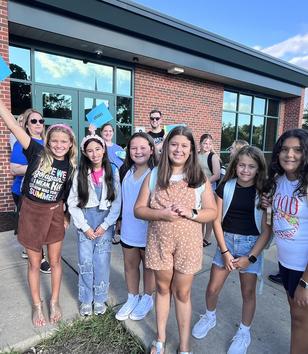 A group of smiling children and adults gathered outside a building, holding signs and posing.