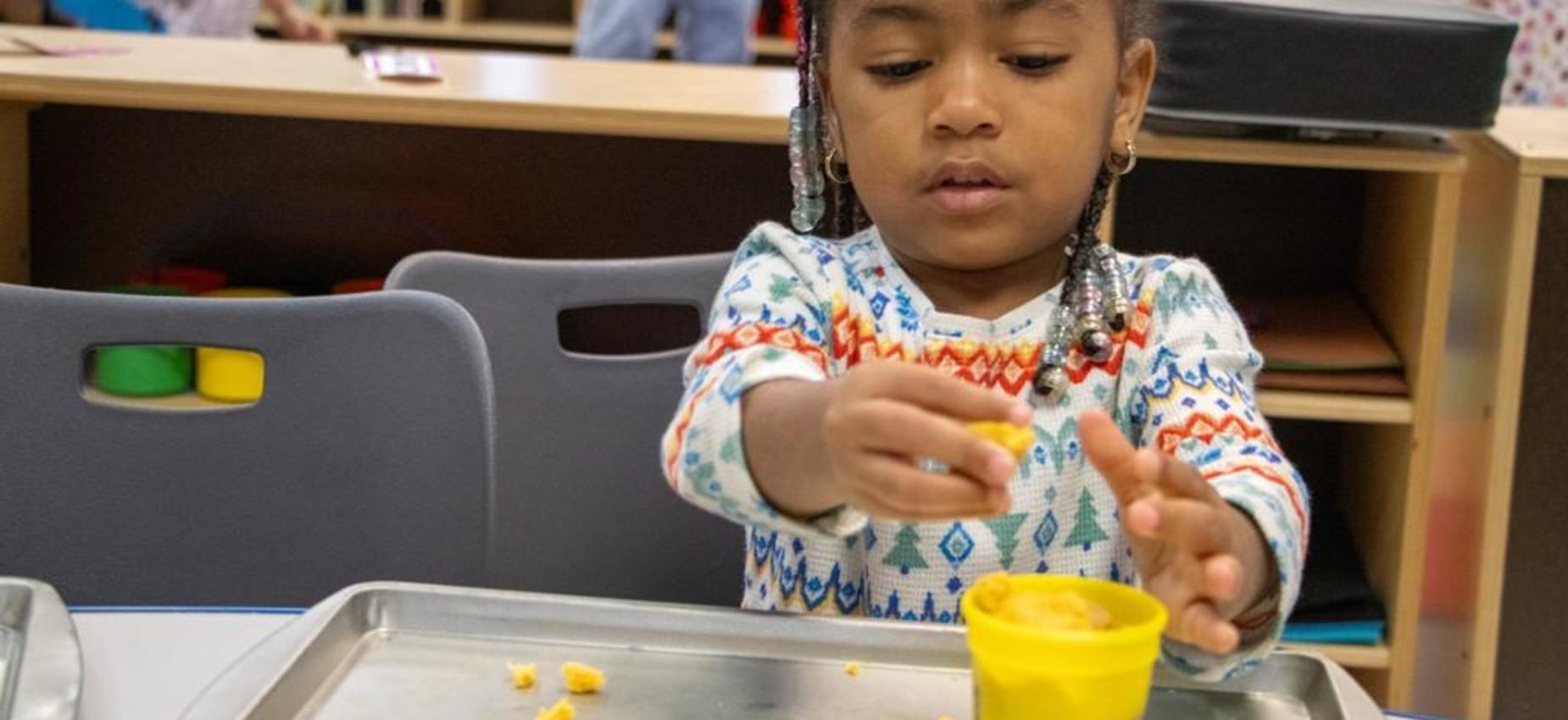 Child playing with playdough while sitting at a table in a classroom.