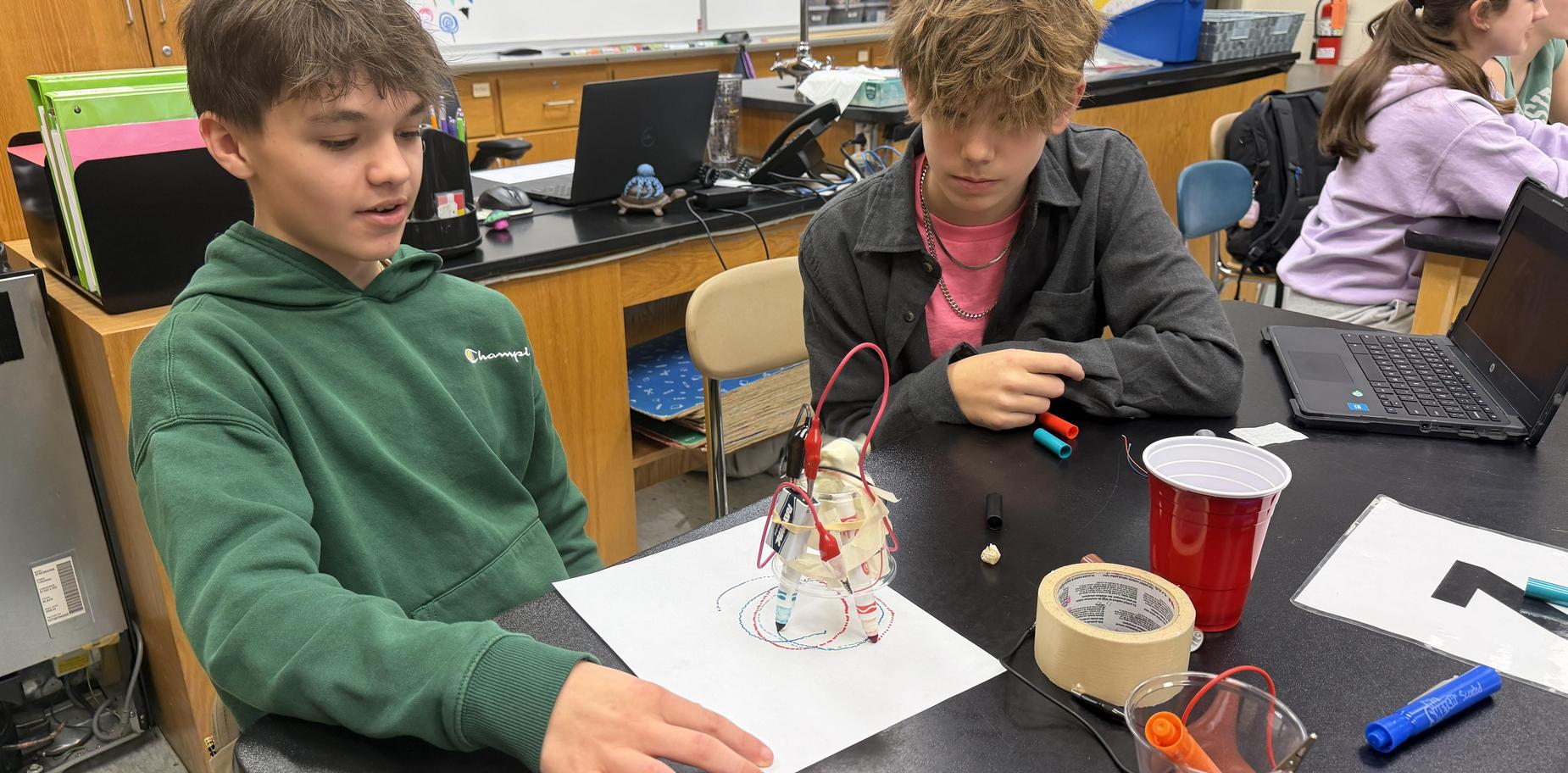 two boys sit at science table watching a robot they made draw on paper