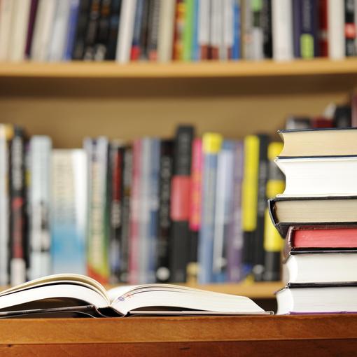 Open book on a wooden table with stacked books and a blurred bookshelf in the background.
