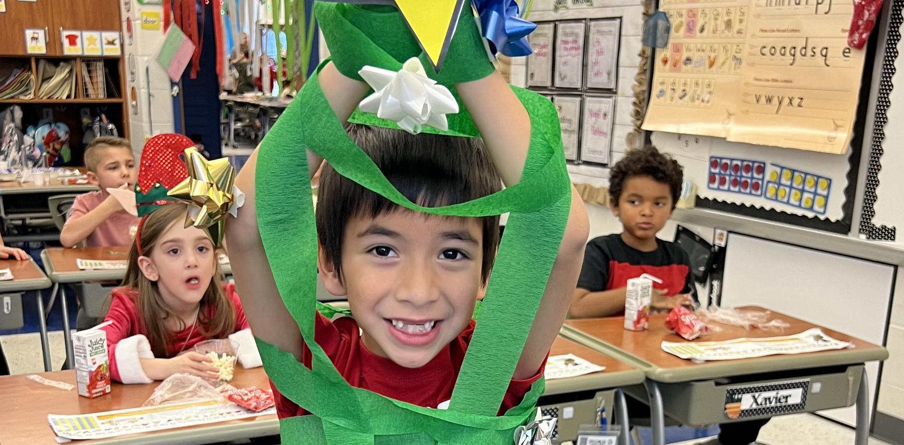 Child in classroom wearing green streamers as a Christmas tree, smiling widely.