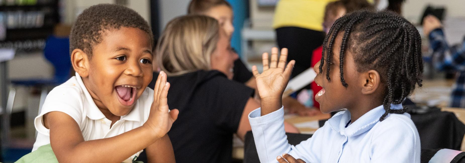 Two kindergarteners wave at each other while seated at a table in a classroom